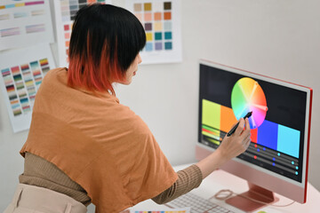 Side view of asian female graphic designer choosing colors from color swatch samples on computer display in creative office