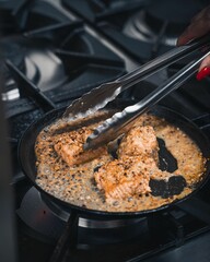 Close up of tongs in a woman's hand cooking sizzling salmon in a pan on the stove