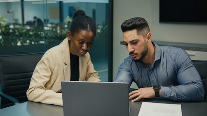Diverse Indian Arabian man and African American woman colleagues discuss financial data analyzing documents marketing project result in report use laptop talk business conversation sit at office desk