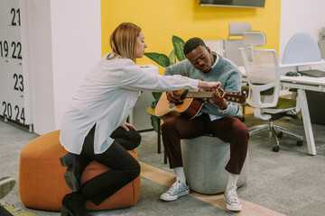 A blonde female person teaching her black male coworker how to play the song on guitar correctly