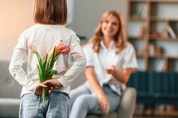 Girl is giving flowers for a woman, holidays. Mother with her little daughter is together in domestic room