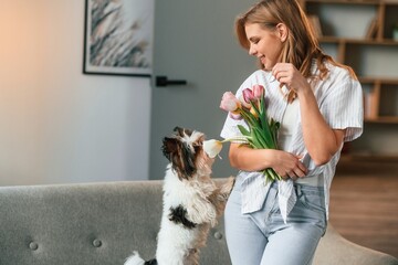 With flowers in hands. Beautiful young woman is with little dog indoors