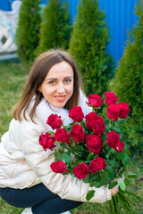 A girl in a white jacket crouched down with a bouquet of red roses near green thuja trees.