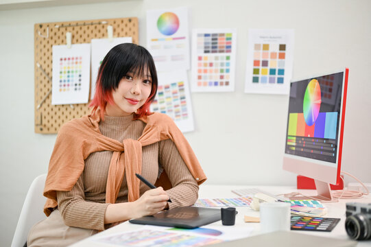 Smiling Asian female graphic designer or interior designer sits at her desk in her studio.