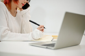 Close-up image of a young Asian woman taking notes while talking on the phone