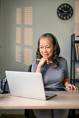 Asian-aged businesswoman working on her business tasks on laptop in her private office.
