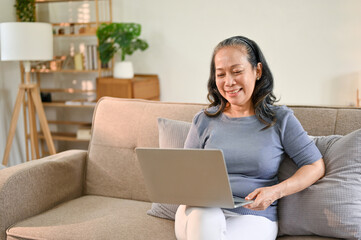 Cheerful Asian-aged retired woman enjoys watching movie on her laptop in her living room