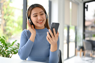 Happy Asian woman sits at a table, chooses a music playlist on her phone, listens music