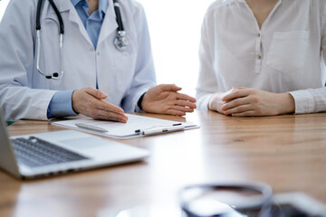 Doctor and patient discussing something while sitting near each other at the wooden desk in clinic, close up of hands. Medicine concept