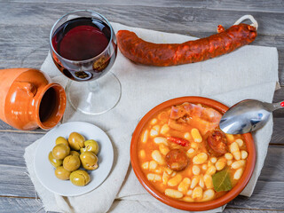 white beans stewed with bacon and chorizo in an earthenware casserole on a wooden table with all the ingredients of the recipe