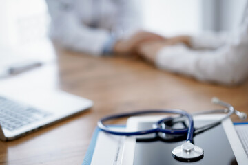 Stethoscope, tablet and laptop computer are lying on the wooden table while doctor's hands reassuring a patient at the background. Medicine concept