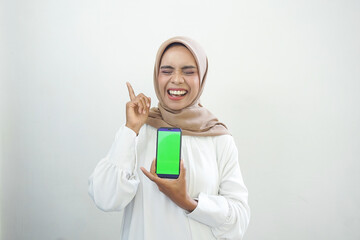 Excited beautiful Asian muslim woman showing green screen mobile phone isolated over white background.