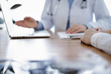 Doctor and patient sitting at the wooden desk in clinic. Female physician's hands pointing into laptop computer monitor, close up. Medicine concept