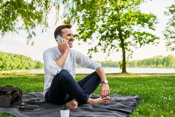 Fototapeta na wymiar Confident mid adult man talking on mobile phone in park sitting on picnic blanket
