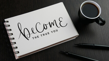 BECOME THE TRUE YOU black hand lettering in notebook with cup of espresso and pens on black wooden desk