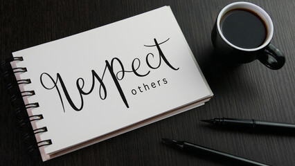 RESPECT OTHERS black hand lettering in notebook with cup of espresso and pens on black wooden desk