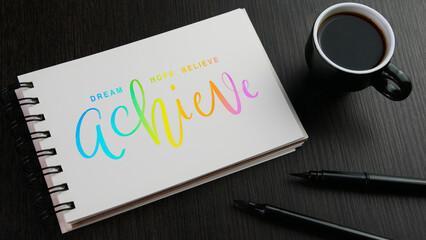DREAM HOPE BELIEVE ACHIEVE colorful hand lettering in notebook with cup of espresso and pens on black wooden desk