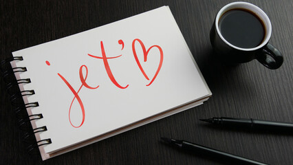 JE T'AIME (I LOVE YOU in French) red hand lettering in notebook with cup of espresso and pens on black wooden desk