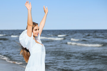 Fototapeta na wymiar Happy smiling beautiful woman on the ocean beach standing in a white summer dress, raising hands