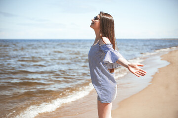 Fototapeta na wymiar Happy smiling woman in free bliss on ocean beach standing with open hands. Portrait of a brunette female model in summer dress enjoying nature during travel holidays vacation outdoors