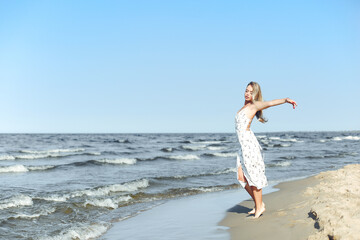 Happy blonde beautiful woman on the ocean beach standing in a white summer dress, open arms