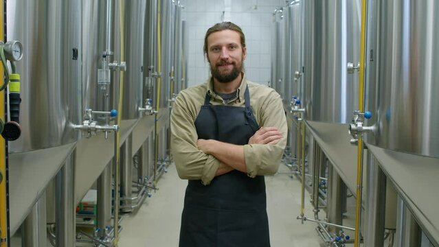 Medium portrait shot of cheerful bearded young Caucasian male technician in apron standing at his workplace in craft brewery, crossing arms on chest, looking at camera and smiling confidently