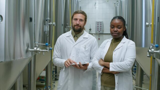 Medium portrait shot of Caucasian male and African American female technologists in white coats standing at work in brewery, next to fermenters and bright beer tanks, looking at camera and smiling