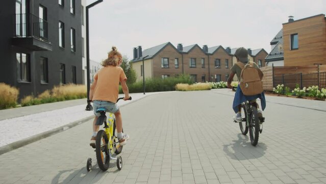 Full rear shot of two 9-year-old children in casual clothes pedaling on bicycles with training wheels on paved street of modern suburban housing estate on sunny summer weekend