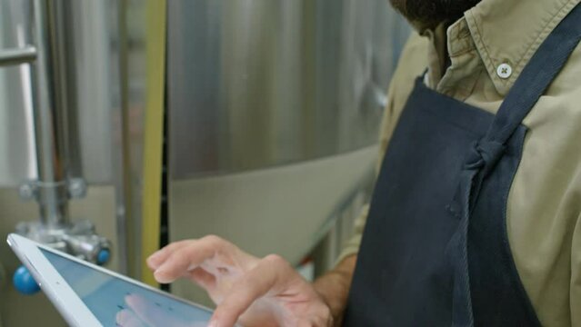 Close-up tilting shot of hands and face of young Caucasian male brewmaster with red hair and beard standing near beer fermentation tank, working on touchscreen tablet and looking at control gauges