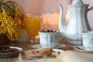 Cup of tea or coffee, plate of cookies, cup of blueberries, plate of chocolate, glass of juice, book, reading glasses, teapot, flowers and lit candles on the table. Brunch or afternoon tea concept. 