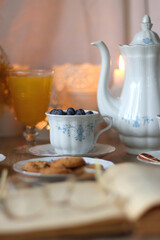 Cup of tea or coffee, plate of cookies, cup of blueberries, plate of chocolate, glass of juice, book, reading glasses, teapot, flowers and lit candles on the table. Brunch or afternoon tea concept. 