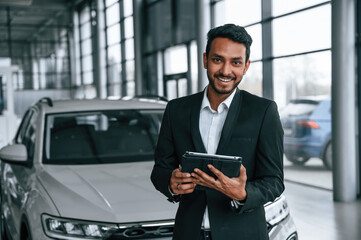 Holding graphic tablet. Handsome indian man in suit is in the car showroom with the vehicle indoors