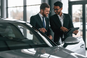 New plan. Standing near automobile with graphic tablet. Two businessmen are working together in the car showroom