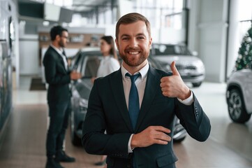 Handsome man is standing in front of his coworkers. Three people are together in the car showroom