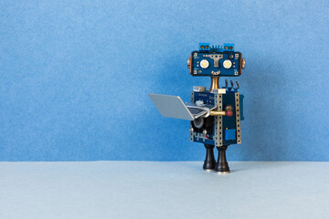 Toy robot bot holds an open compact silver laptop. Modern artificial intelligence machine learning technologies. Copy space on blue background - 587267364
