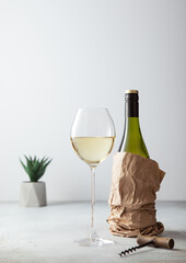 Bottle of white wine in paper bag with opener and glass of wine on light background.