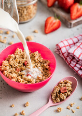 Pouring milk into bowl with strawberry granola on light kitchen board.