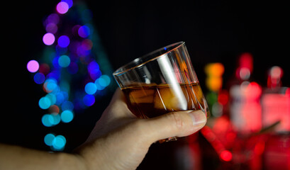 A glass with an alcoholic drink in hand against the background of the bar. Whiskey with ice in hand.