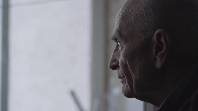 Elderly person looks out window at home and enjoys spending quiet life. Portrait of lonely old wrinkled man thinking about future life