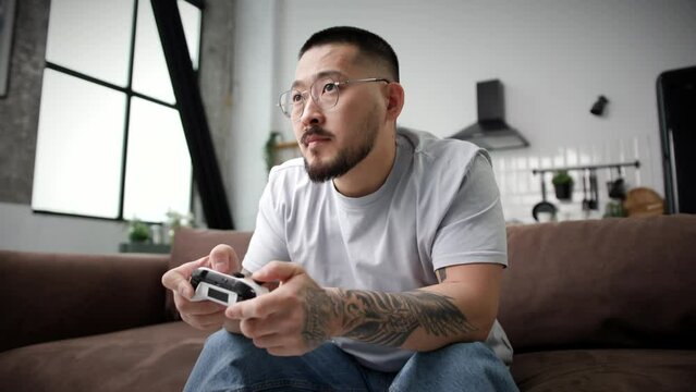 Man Playing Video Game Holding Joystick. Asian man playing games with controller sitting on sofa at home. Handheld Camera.