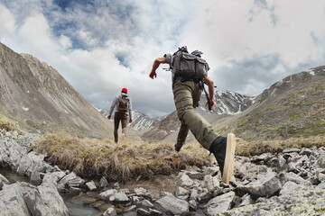 Two young hikers walks with light backpacks in mountains. Tourist jumps across the obstacle - obrazy, fototapety, plakaty