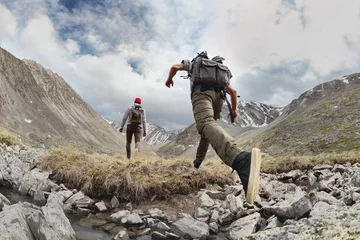 Fotobehang Two young hikers walks with light backpacks in mountains. Tourist jumps across the obstacle © cppzone