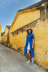 Backpacker woman in blue dress stands on a yellow wall. Tourists walking in Hoi An old town Vietnam