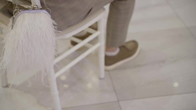 Man in free form brown business suit sits on white chiavari chair with elegant white pouch with feathers. Inside hall with white tiles on floor, brown casual shoes with white short socks.