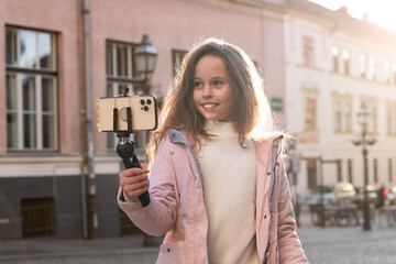 Young happy preteen girl recording a vlog outside in a sunny day, wearing a pink jacket, being online