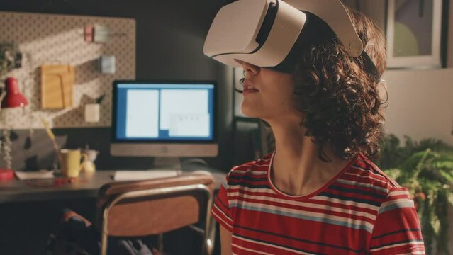 Tilt up shot of curly young girl pressing space on laptop, wearing white VR headset, bright T-shirt, shorts and sneakers, sitting on bed in morning, moving her head. Desk and monitor screen behind
