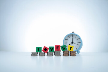 The word INVEST is written on the wooden cubes on the stacked coins. An alarm clock at the back. Time to Invest concept