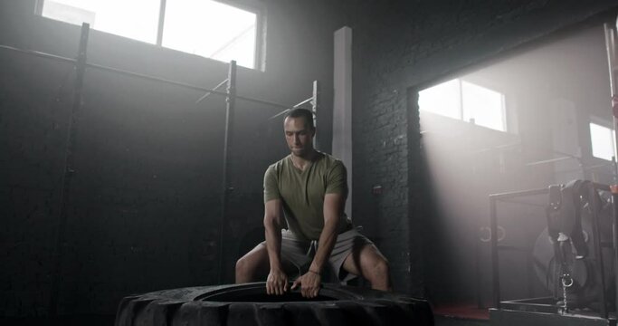 Handsome African American strongman training in shadow sports hall. Attractive sportsman lifting, flipping heavy, huge wheel or rubber tyre. Guy pumping muscles and bodybuilding. Powerlifting concept