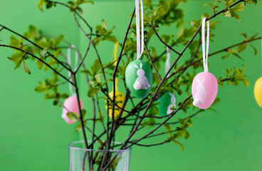 Colorful Easter eggs with cute white bunnies hanging on green tree branches in glass vase, green background. Easter concept. Easter still life. Closeup of Easter home decoration.