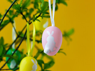 Colorful Easter eggs with cute white Easter bunnies hanging on green tree branches, yellow background. Easter concept. Easter still life. Closeup of Easter home decoration.